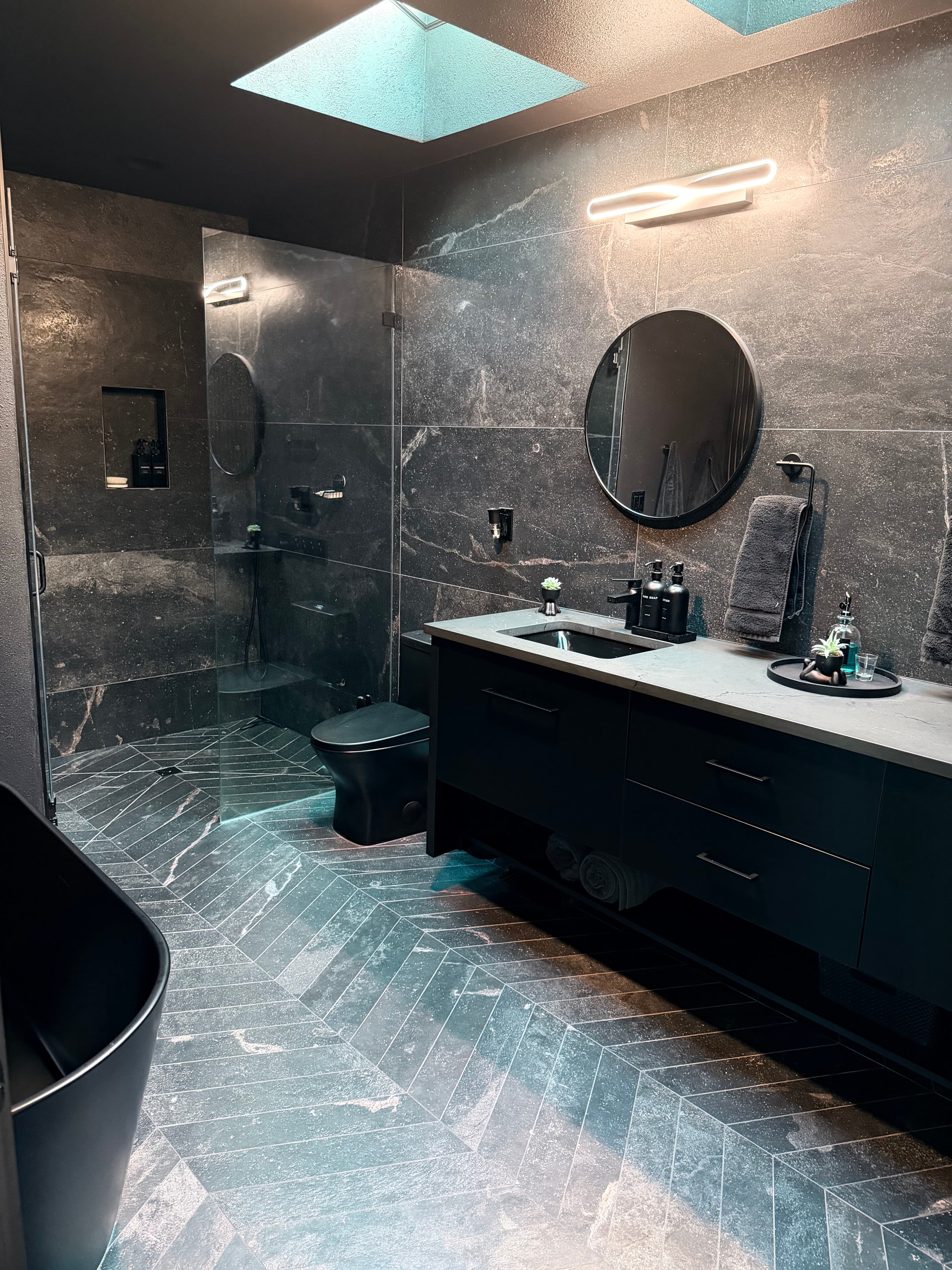 Bathroom remodel in dark marble-look tile with a round mirror, black fixtures, and a skylight